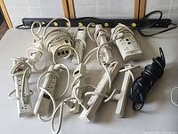 Photo showing multiple white power strips with varying numbers of outlets and USB charging ports, along with a long black power strip on a beige surface.