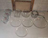 Clear glass Pyrex measuring cups, baking dishes, pie plates, and saucepan with lid arranged on a white surface with folding chairs in background.