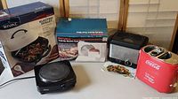 Overall view of four small kitchen appliances: Rival Electric Skillet, Rival Folding Blade Food Slicer, Oster Ceramic Hot Plate, Coca-Cola electric can cooler