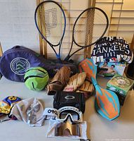 Mixed sports equipment including two tennis rackets with a cover, multiple baseball gloves, a small soccer ball, skate shoes, Oakley goggles, and sports apparel.