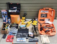 Overall view of lot including cordless tools, battery packs, tool kits, safety gear, and storage items laid out on concrete floor.