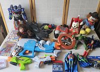 Front view of lot showing robot action figure, plush monkey, toy guns, clown doll, dog plush, and action figures laid out on table with comics and games