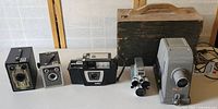 Photo showing five vintage photographic items including three cameras, an 8mm film camera, and a Bell & Howell slide projector with wooden case.