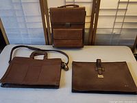 Photo of three vintage brown leather attache cases, one standing upright with multiple front pockets and lock, one slim case with handle and shoulder strap laying flat, and one Zackly Farms folio with clasp and lettering.