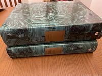 Two hardcover volumes with green marbled covers stacked on a table, displaying the gold title plaques on their spines.