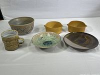 Set of six ceramic pottery items including two mustard yellow handled bowls, one large bowl with branch motif, a ribbed mug, and two plates, displayed on white surface