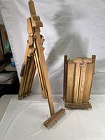 Two wooden portable easels shown side by side, one set up and one folded flat for storage with natural wood and brass hardware visible.