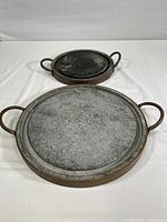 Two copper and soapstone griddles shown together on a white surface; larger griddle in front with light gray surface and copper handles; smaller griddle behind with darker surface and copper handles