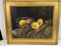 Framed oil painting depicting a still life of fruit in a clear glass bowl with a dark background and colorful draped cloth beneath.