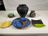 Group view of the pottery and ceramic items including the large black embossed vase, abstract plates in blue and green, yellow plate, small brown vase, and green bowl.