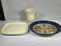 Ceramic pitcher and two dishes, one square white and one round decorative piece displayed on plain background
