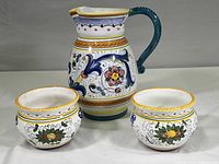 Full view of the ceramic pitcher and two bowls, showing the colorful hand-painted floral and scroll designs on white ceramic with yellow rims and teal handle.