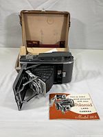 Polaroid Land Camera 110A unfolded with bellows extended, placed in front of open beige carrying case. Manual visible under the camera.