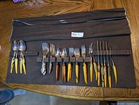 Set of 21 flatware pieces organized on brown fabric roll-up drawer insert, showing spoons, forks, and cocktail forks with yellow Bakelite handles and some stainless steel flatware.