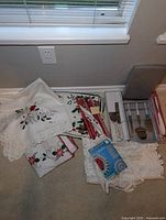 Overview of lace Christmas tablecloths stacked with red candles and boxed cheese knives and cake serving utensils beside them on floor