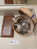 Vintage brown leather wallet with visible internal wear placed beside a round woven basket containing assorted jewelry items including pins, pendants, earrings, and beaded crucifix necklace.