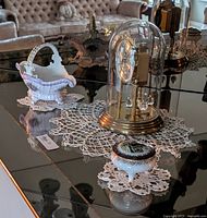 Porcelain basket, lidded trinket dish, and dome clock on a glass table with crocheted doilies beneath each item.