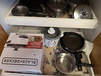 View of kitchen drawer containing several pots and pans with Black+Decker air fryer box, rice cooker, frying pan, and saucepan