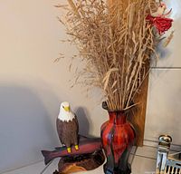 Photo showing red and black pottery vase with dried foliage and wooden bald eagle carving on fish beside it.