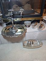 Silver plated candelabra on plated serving platter, crystal salt server, salt and pepper shakers, small crystal dishes with forks arranged on a tiled hearth in front of a fireplace.