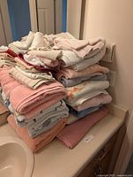 Stack of assorted towels and handcloths folded on a bathroom counter next to a sink and electrical outlet.