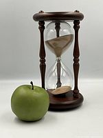 Full view of the vintage wooden sand timer hourglass alongside a green apple for size reference. The hourglass is 12 inches tall with a wooden frame and clear glass bulbs containing light brown sand.