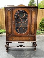 Front view of the 1920's walnut cabinet showing the glass door with intricate wood mullions, drawer, and base with turned legs.