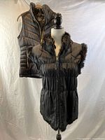 Both vests shown together on mannequin and hanger: black Michael Kors down vest and black wool vest with fur trim.