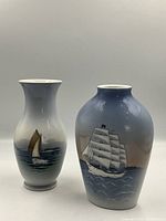Front view of two Royal Copenhagen vases, one with a ship painting and one with a sailboat painting, both standing upright on neutral background.