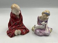 Front view of two Royal Doulton figurines: A boy wrapped in a red blanket seated and a girl in a purple dress with a lamb on lap.