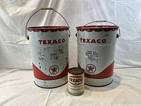 Three Texaco cans: two large 16-inch empty oil cans and one small full paint can 6 inches high, front view showing branding and condition.