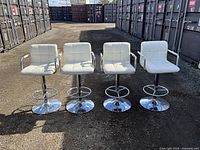 Four white leather bar stools with chrome bases lined up outdoors, showing front view and armrests.