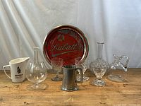 Photo of vintage Labatt's beer tin litho tray, glass decanters, brandy balloon glass, pewter tankard, Dewar's pitcher, and wine glasses on wooden surface against light backdrop.