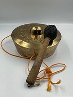 Brass gong with leather-bound striker placed on white surface, showing side view with orange cord attached.