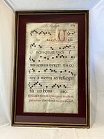 Framed manuscript sheet showing illuminated initial and neumatic notation, red mat border and gold frame on white background