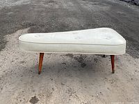 Front view of a white triangular mid century modern bench or ottoman with three teak legs and brass caps on legs.