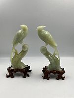 Pair of jade bird sculptures on stands against neutral background