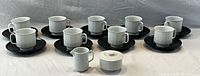 Full view of 9 white porcelain tea cups on black saucers, with creamer and sugar bowl at front, all with vertical ribbed pattern and mid-century modern design.