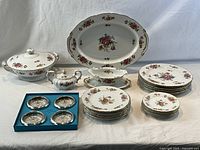 Full set of SANGO Dresdenia porcelain china displayed with covered dish, large platter, multiple plates of various sizes, gravy boat with tray, lidded sugar bowl, and Birks silver plate coasters in original box. All items arranged on table for clear view of pattern and quantity.