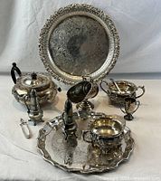 Full set of silver plated and pewter tableware including trays, teapot, sugar bowl with spoon, shakers, caster, trophy cup and tongs arranged on white cloth background.