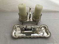 Front view of the silver plated double candle holder with two half-white pillar candles and the matching snuffer/trimmer on the tray