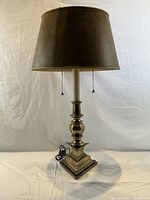 Solid brass table lamp with original dark fabric shade, shown lit against white backdrop.