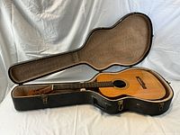 Open black hard guitar case with plush tan lining housing a natural wood finish acoustic guitar.