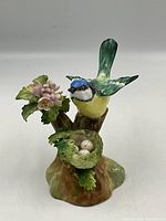 Porcelain figurine of a Tom Tit bird on a branch with flowers and a nest with eggs, photographed against a plain background.