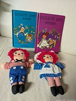 Photo showing two 1989 Raggedy Ann and Raggedy Andy plush dolls with their two vintage storybooks displayed upright behind them on a white couch.