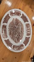 Top-down view of white oval porcelain meat plate with brown engraved historical maritime scenes and decorative borders.