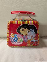 Front view of mini metal lunchbox showing Dora hugging Boots with Nick Jr. Dora the Explorer logo and striped border around edges.