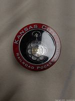Pocket watch displayed inside original round tin case with clear front window and red border marked 'Kansas City Railroad Pocket Watch'. Watch has Roman numeral dial and visible gears.