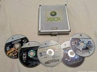 Five Xbox 360 game discs laid out with a silver metal Xbox-branded multi disc holder on a beige fabric surface.