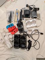 Full view of assorted gaming controllers and accessories including Nintendo Switch Joy-Con, Wii remotes, chargers, and cables on a beige surface.